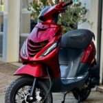 Rode scooter met schijfrem en LED-koplampen, geparkeerd voor een gebouw.