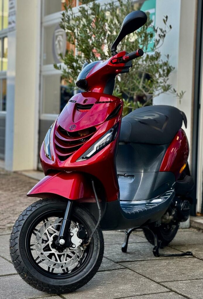 Rode scooter met schijfrem en LED-koplampen, geparkeerd voor een gebouw.