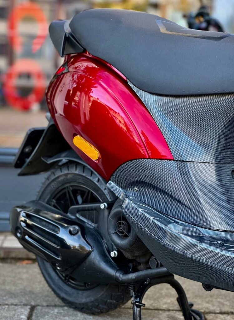 Close-up van de achterzijde van een rode scooter met zwart zadel, achterwiel en uitlaat.