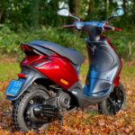 Rood-zwarte scooter geparkeerd op herfstbladeren aan de bosrand, achterdriekwart aanzicht met spiegels.