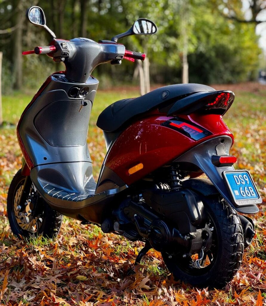 Rood-zwarte scooter geparkeerd op herfstbladeren in een park met bomen op de achtergrond.