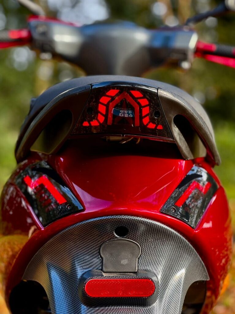 Close-up van de achterzijde van een rode scooter met rode LED-achterlichten en carbonlook panelen.