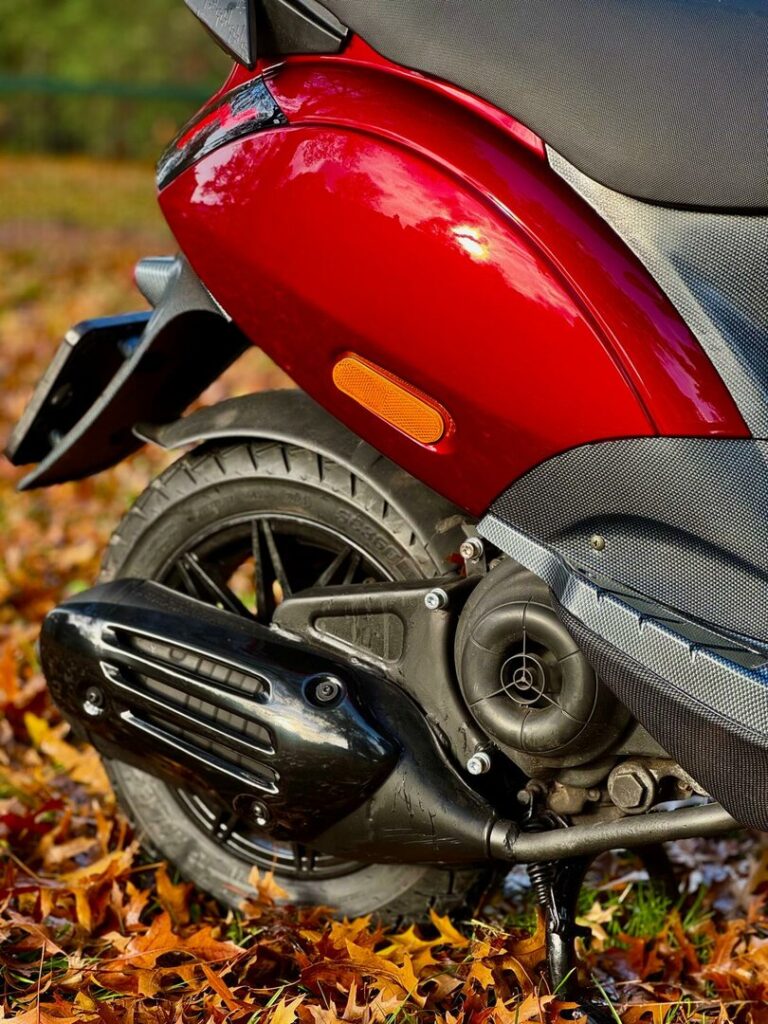 Close-up van de achterzijde van een rode scooter met wiel en uitlaat op herfstbladeren.