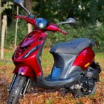 Rode scooter geparkeerd op herfstbladeren in een groen bos, met koplamp aan.