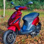 Glanzend rode scooter geparkeerd op herfstbladeren, met bos op de achtergrond.