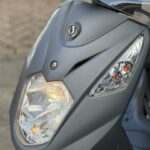 Close-up van de voorzijde van een grijze scooter met brandende koplamp en ruit.