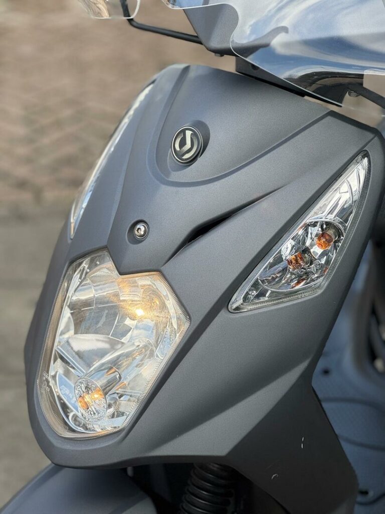 Close-up van de voorzijde van een grijze scooter met brandende koplamp en ruit.