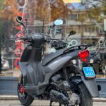 Matgrijze scooter met hoog windscherm op de standaard, voor een winkelraam met herfstreflectie.