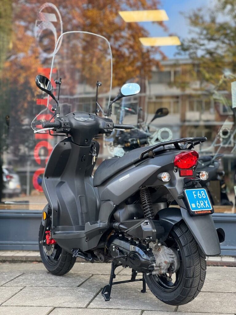 Matgrijze scooter met hoog windscherm op de standaard, voor een winkelraam met herfstreflectie.