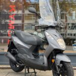 Grijze scooter met hoog windscherm op standaard, geparkeerd voor een winkelraam.