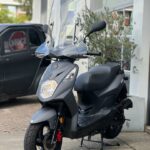 Matgrijze motorscooter met hoog windscherm op standaard, geparkeerd bij een gebouw.