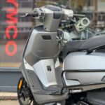 Close-up van een grijze scooter met spiegels, geparkeerd voor een winkel met KYMCO-logo.