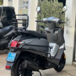 Matgrijze scooter met achterdrager en spiegels, geparkeerd voor een werkplaats, achteraanzicht.