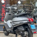 Grijze Kymco Like scooter op standaard, met achterdrager en blauwe kentekenplaat, voor een winkelraam.