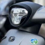 Close-up van de koplamp en voorkap van een matte grijze scooter met E4-sticker.