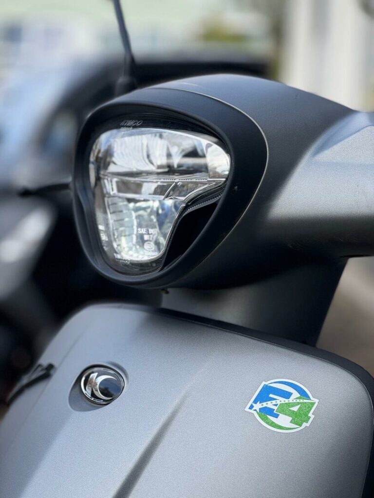 Close-up van de koplamp en voorkap van een matte grijze scooter met E4-sticker.