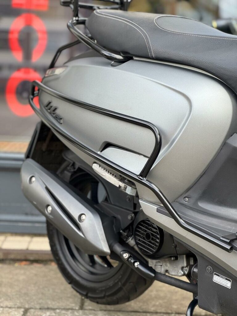 Close-up van de achterzijde van een grijze scooter met uitlaat, valbeugels en zwart zadel.