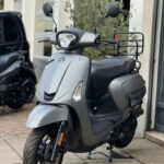 Matgrijze stadscooter met spiegels en voorrek, geparkeerd bij een garage.