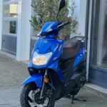 Blauwe SYM-scooter op zijstandaard voor een winkel, frontaal gezien.