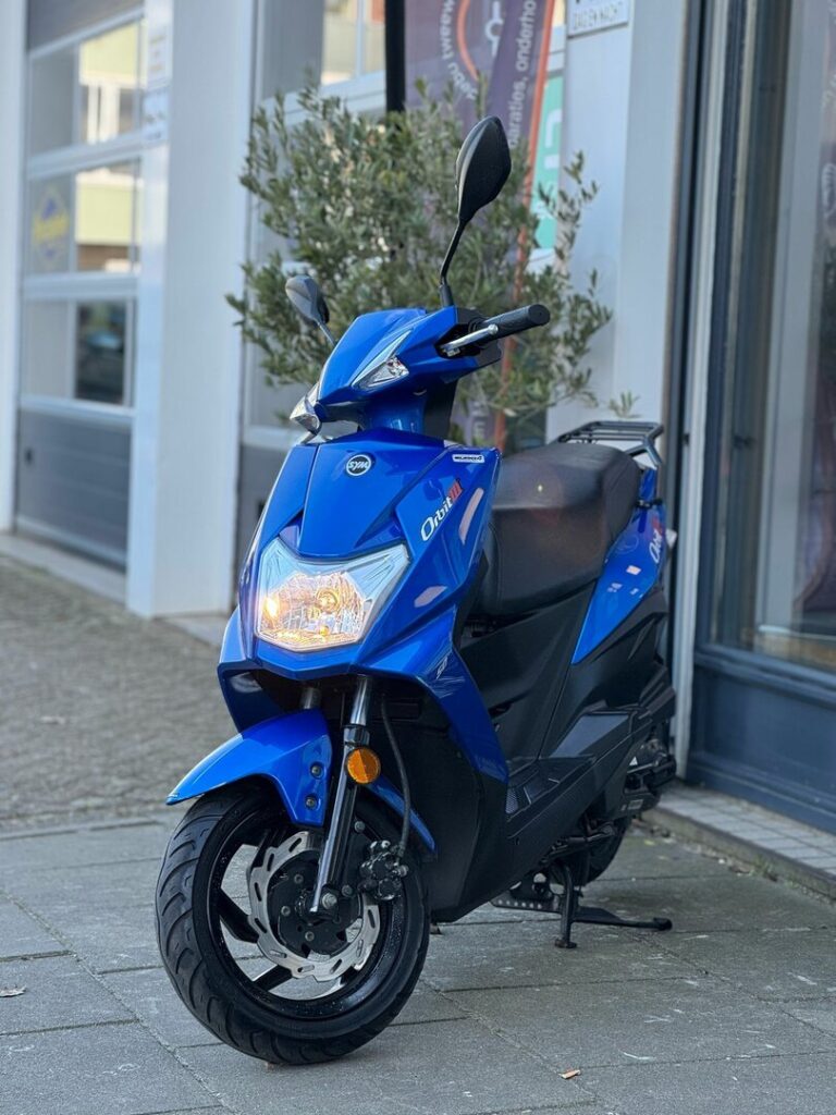 Blauwe SYM-scooter op zijstandaard voor een winkel, frontaal gezien.