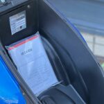 Onder het zadel van een scooter: zwart opbergvak met documentmap en typeplaatje.