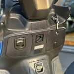 Close-up van het stuur en de middenconsole van een scooter met contactsleutel en opbergvakken.