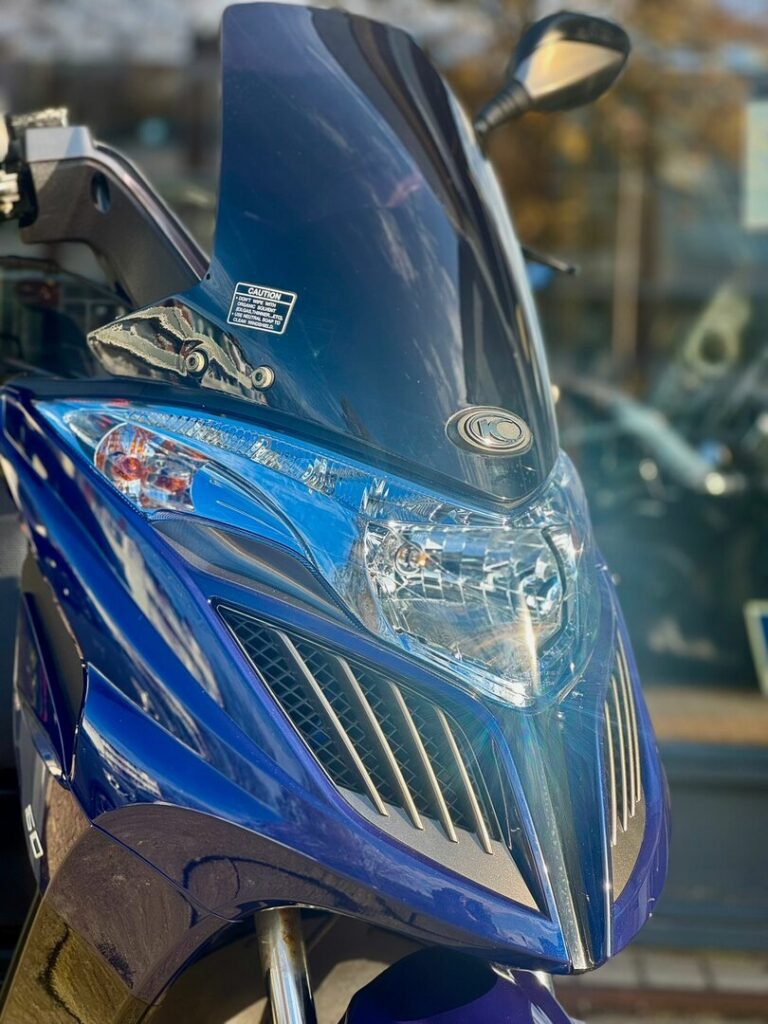 Close-up van de voorkant van een blauwe scooter met hoog windscherm en koplamp.