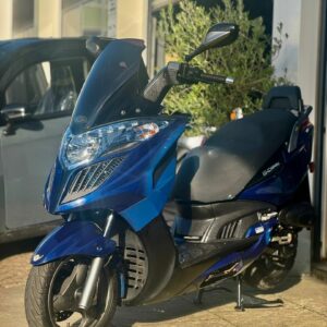 Blauwe motorscooter op standaard voor een garage, in zonlicht op trottoir.