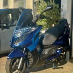 Blauwe motorscooter geparkeerd voor een garage, in de zon, met grote koplampen en windscherm.