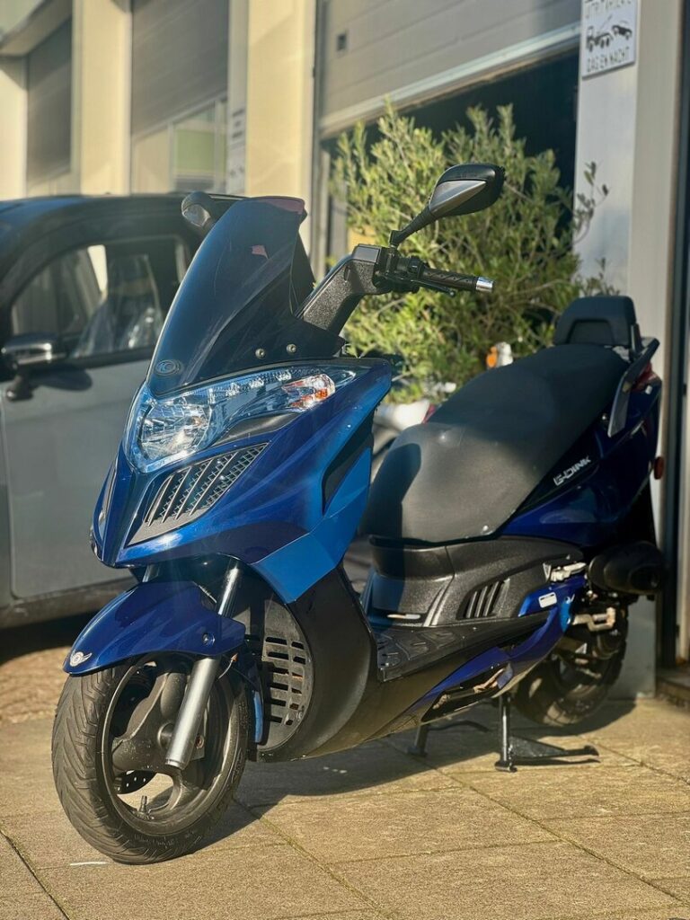 Blauwe motorscooter geparkeerd voor een garage, in de zon, met grote koplampen en windscherm.