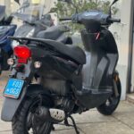 Zwarte scooter op standaard met blauwe kentekenplaat, achteraanzicht op stoep bij winkel.