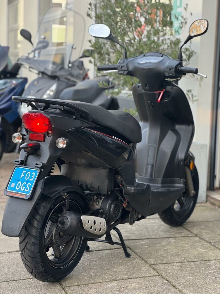 Zwarte scooter op standaard met blauwe kentekenplaat, achteraanzicht op stoep bij winkel.
