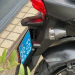 Close-up van de achterzijde van een scooter met rood achterlicht en blauw kenteken, geparkeerd op de stoep.