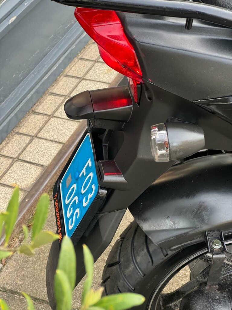 Close-up van de achterzijde van een scooter met rood achterlicht en blauw kenteken, geparkeerd op de stoep.