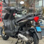 Zwarte scooter op de middenbok, achteraanzicht met blauwe kentekenplaat, geparkeerd voor een winkelraam.