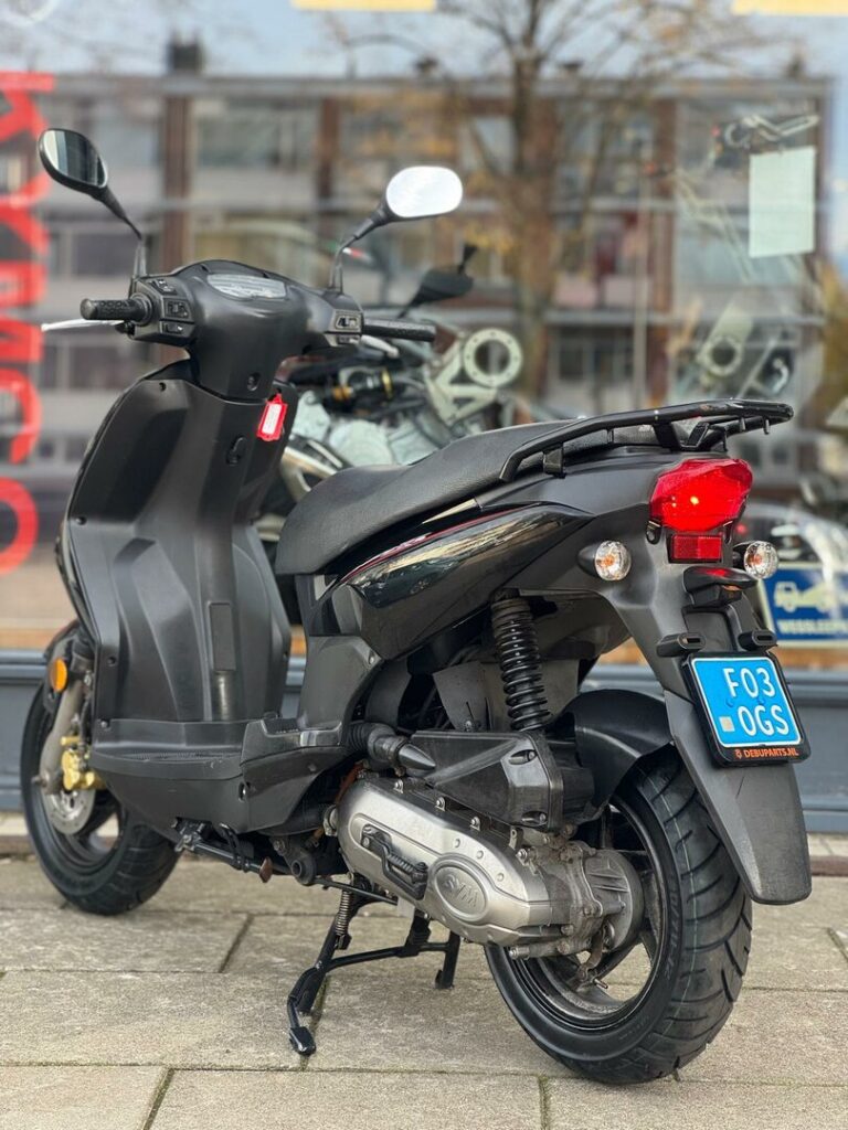 Zwarte scooter op de middenbok, achteraanzicht met blauwe kentekenplaat, geparkeerd voor een winkelraam.