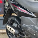 Achterzijde van een zwarte scooter met krassen op de kuip en uitlaat, geparkeerd op de stoep.