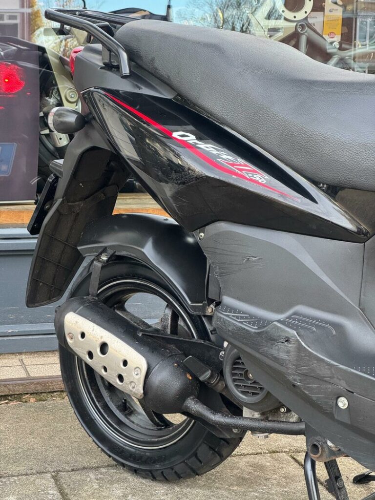 Achterzijde van een zwarte scooter met krassen op de kuip en uitlaat, geparkeerd op de stoep.