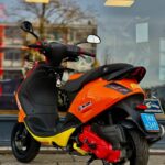 Oranje-gele scooter met rode accenten op standaard, geparkeerd voor een winkelraam.