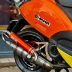 Close-up van oranje scooter met Yasuni uitlaat en MHR-sticker, achterwiel en zadel zichtbaar.