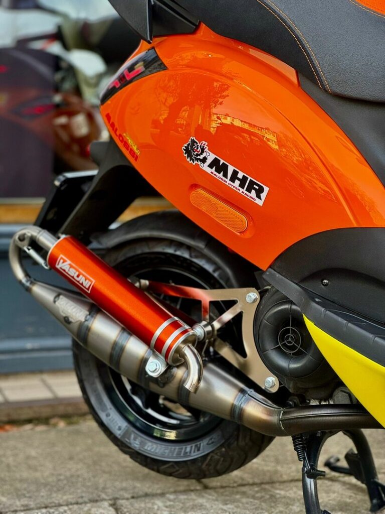 Close-up van oranje scooter met Yasuni uitlaat en MHR-sticker, achterwiel en zadel zichtbaar.