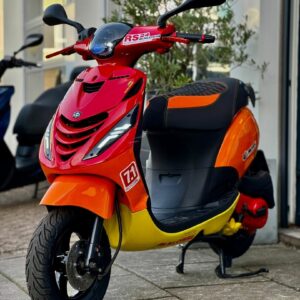 Rood-oranje-gele sportieve scooter met RS24 Malossi-sticker, geparkeerd op de stoep bij een winkel.