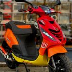 Rood-oranje scooter met sportieve stickers, geparkeerd op trottoir voor winkelraam.
