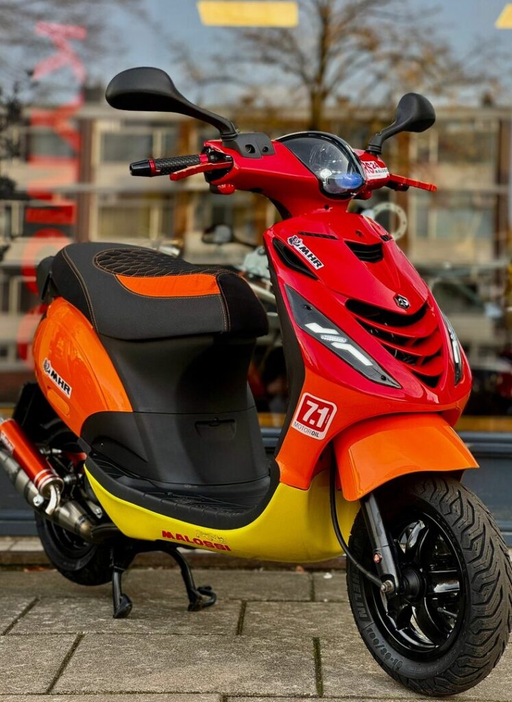 Rood-oranje scooter met sportieve stickers, geparkeerd op trottoir voor winkelraam.
