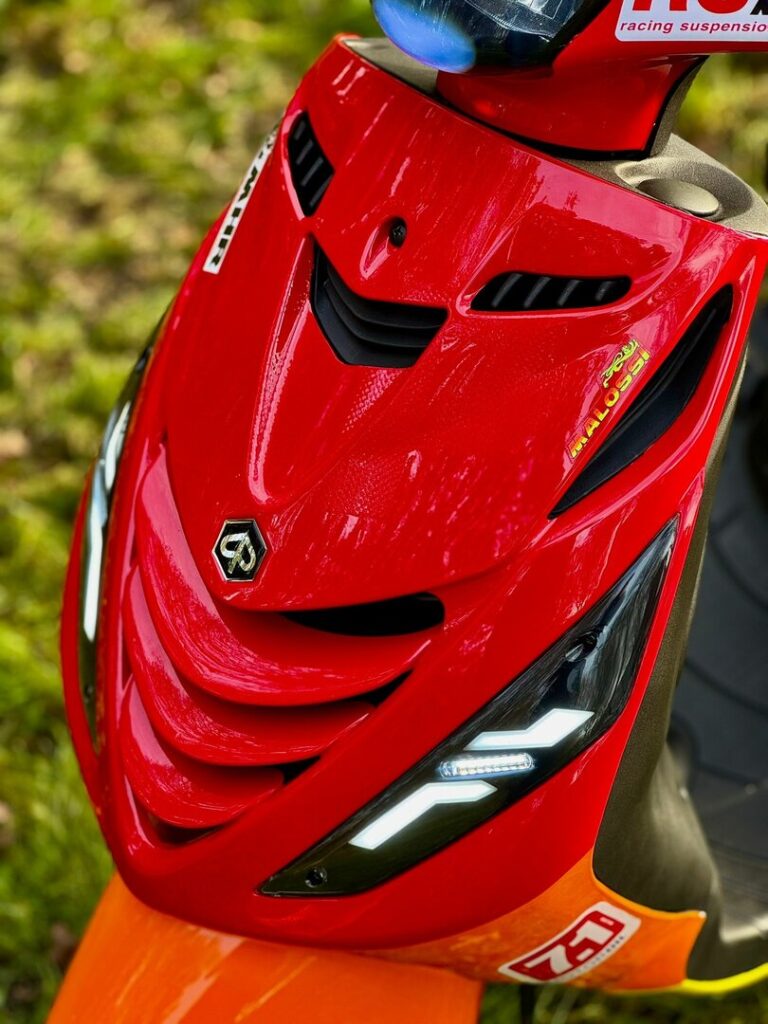 Close-up van de rode voorkap van een sportieve scooter met LED-dagrijlichten en Malossi-sticker.