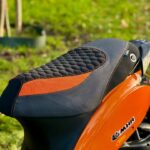 Close-up van een zwart-oranje scootersadel met honingraatstiksel op een oranje MHR-scooter buiten.