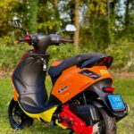 Oranje-gele Malossi-scooter met blauwe kentekenplaat, geparkeerd op gras voor een bosrijke achtergrond.