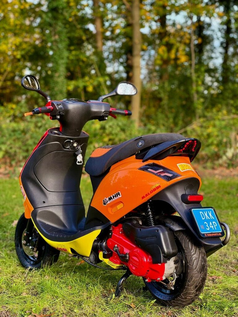 Oranje-gele Malossi-scooter met blauwe kentekenplaat, geparkeerd op gras voor een bosrijke achtergrond.