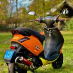 Oranje scooter met sportuitlaat op gras, met bomen en een huisje op de achtergrond.