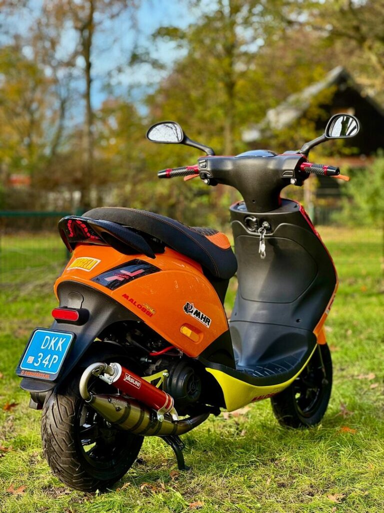 Oranje scooter met sportuitlaat op gras, met bomen en een huisje op de achtergrond.